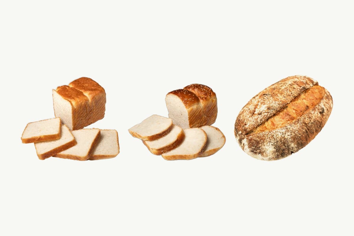 A sliced white toast loaf, a sliced Japanese milk bread loaf and a country sourdough loaf side by side on a plain cream background