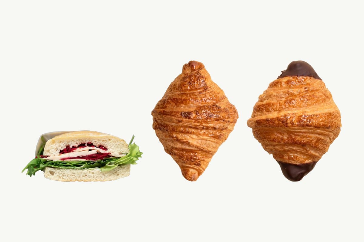 A ciabatta turkey and cheese sandwich, a plain croissant and a chocolate-dipped croissant side by side on a plain cream background