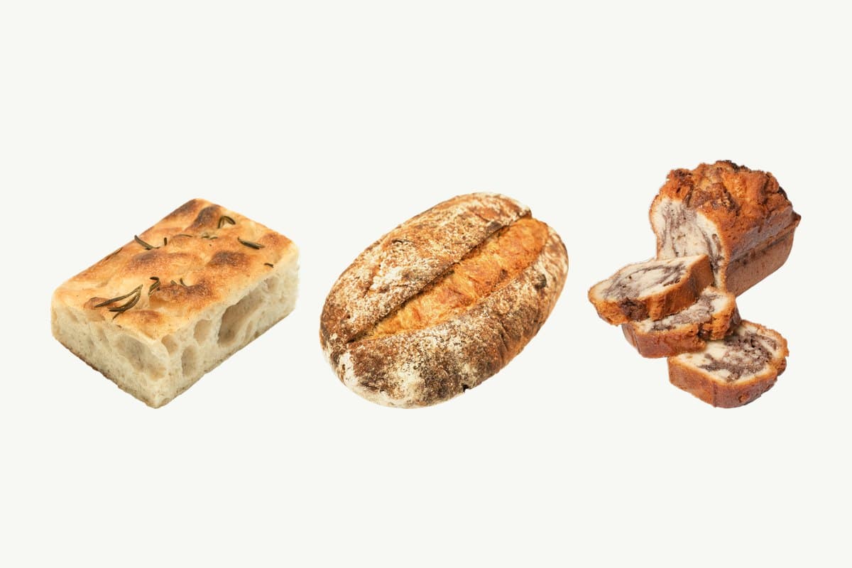 A rosemary and olive oil focaccia, a country sourdough loaf and a sliced marble cake loaf side by side on a plain cream background