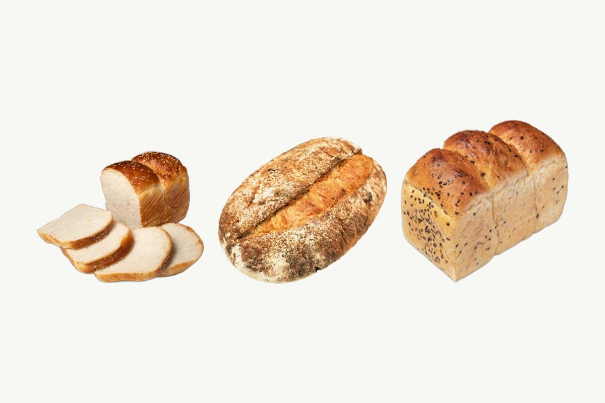A sliced Japanese milk bread loaf, a country sourdough loaf and a multi-seed whole wheat toast loaf side by side on a plain cream background