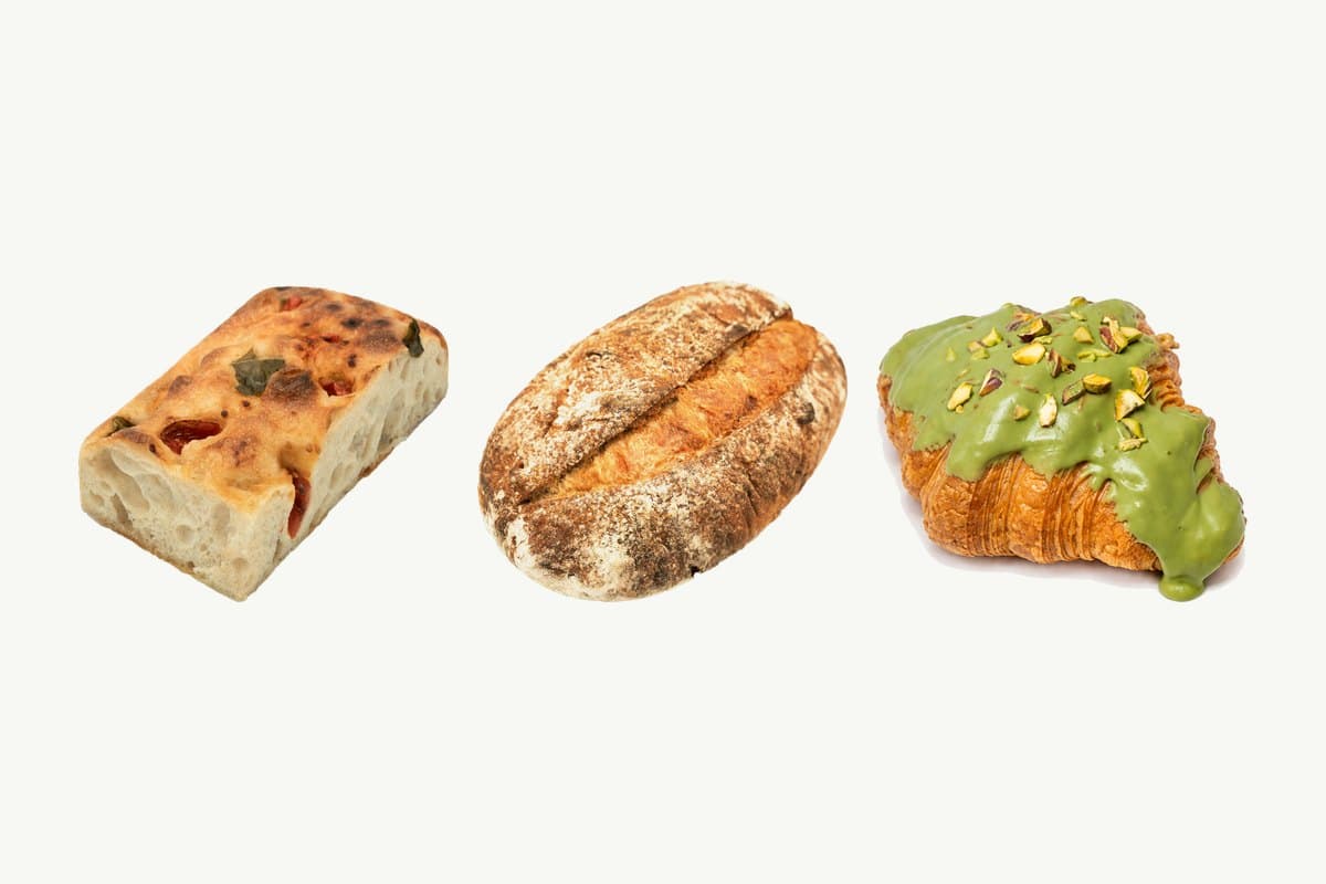 A cherry tomato focaccia, a country sourdough loaf and a pistachio croissant side by side on a plain cream background