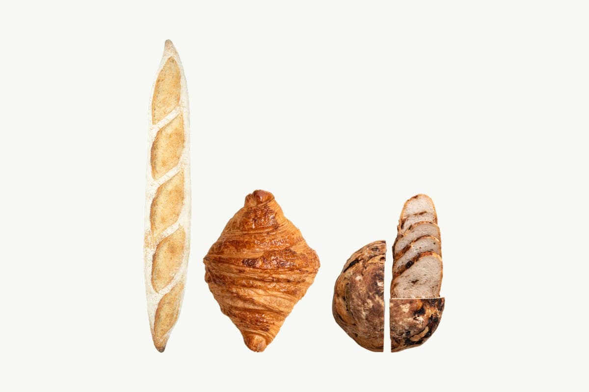 A sourdough baguette, a butter croissant and a Date, Walnut & Honey Sourdough cut open, in a row on a plain cream background