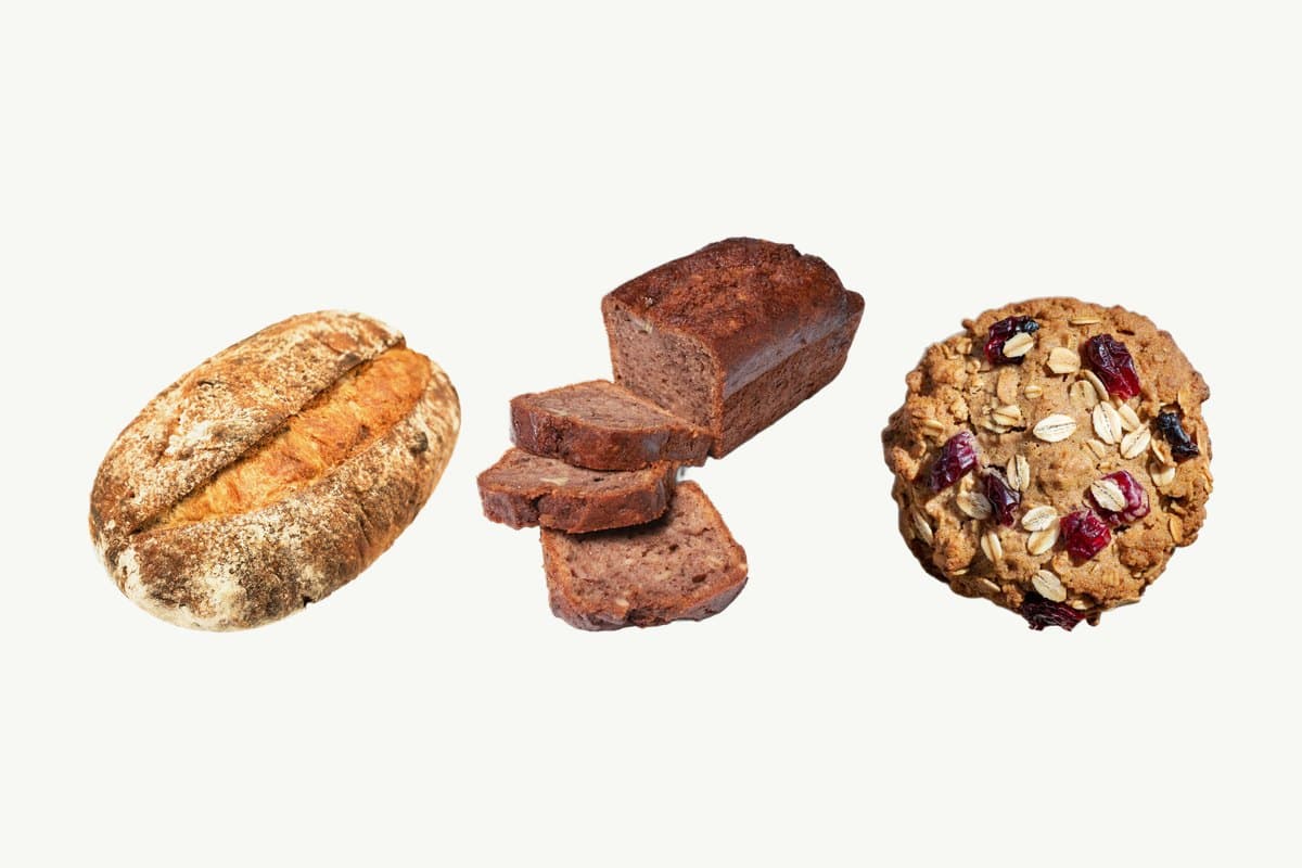 A Country Sourdough Loaf, a partly sliced Banana & Pecan Cake Loaf and an Oats & Cranberry Cookie in a row on a plain cream background