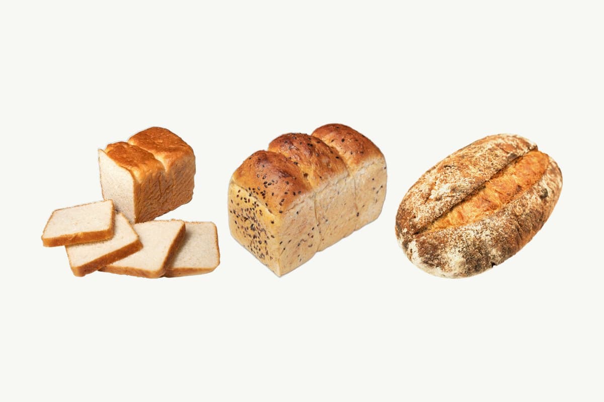 A sliced white toast loaf, a multi-seed whole wheat toast loaf and a country sourdough loaf side by side on a plain cream background