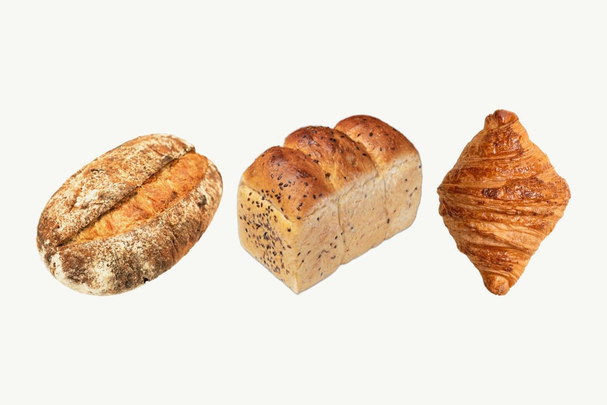 A country sourdough loaf, a multi-seed whole wheat toast loaf and a plain croissant side by side on a plain cream background