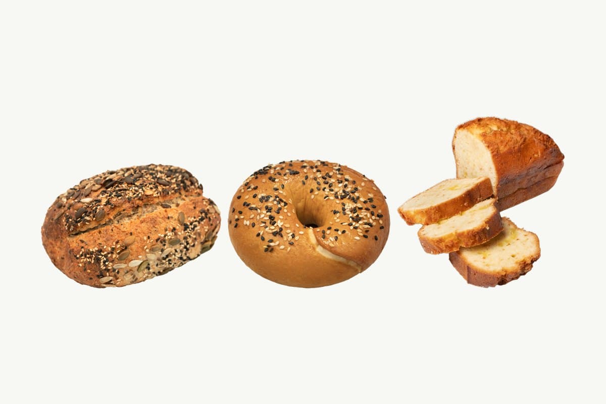 A multi-seed sourdough loaf, a glossy bagel scattered with black and white sesame and a sliced lemon butter cake loaf side by side on a plain cream background