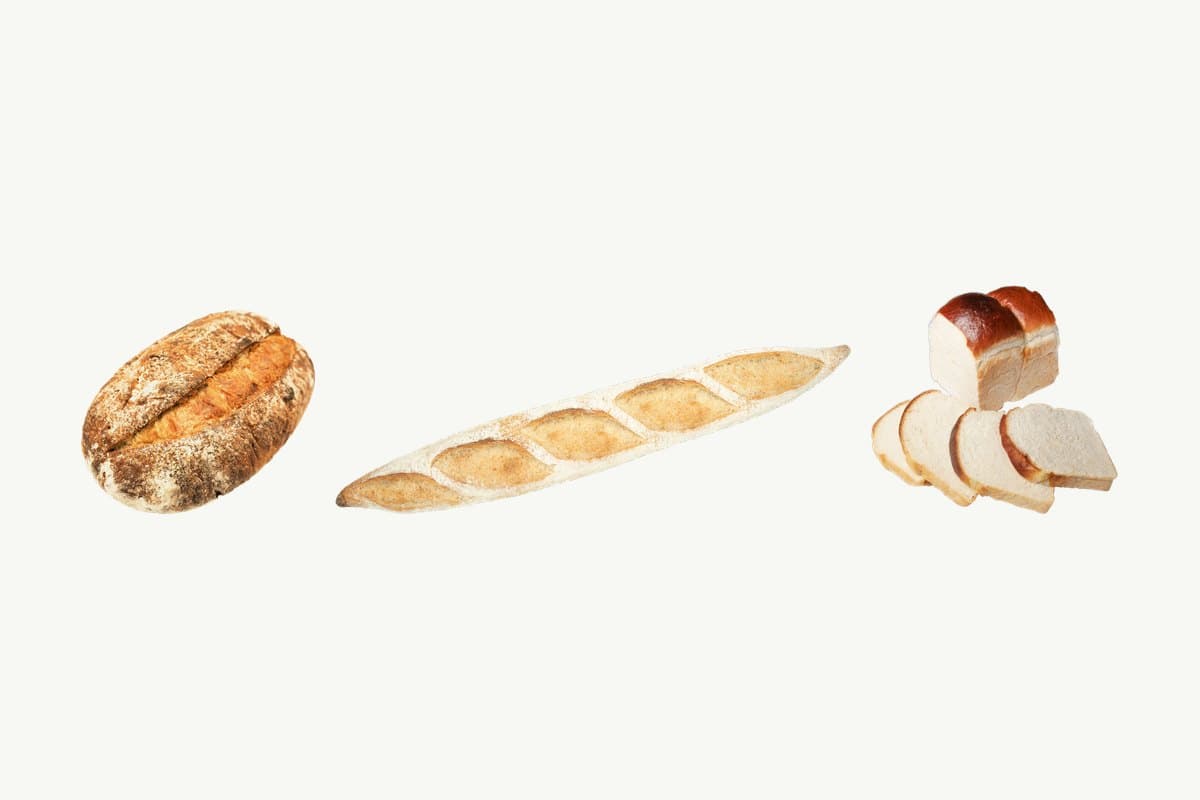 A country sourdough loaf, a sourdough baguette lying on its side and a sliced brioche toast loaf in a row on a plain cream background