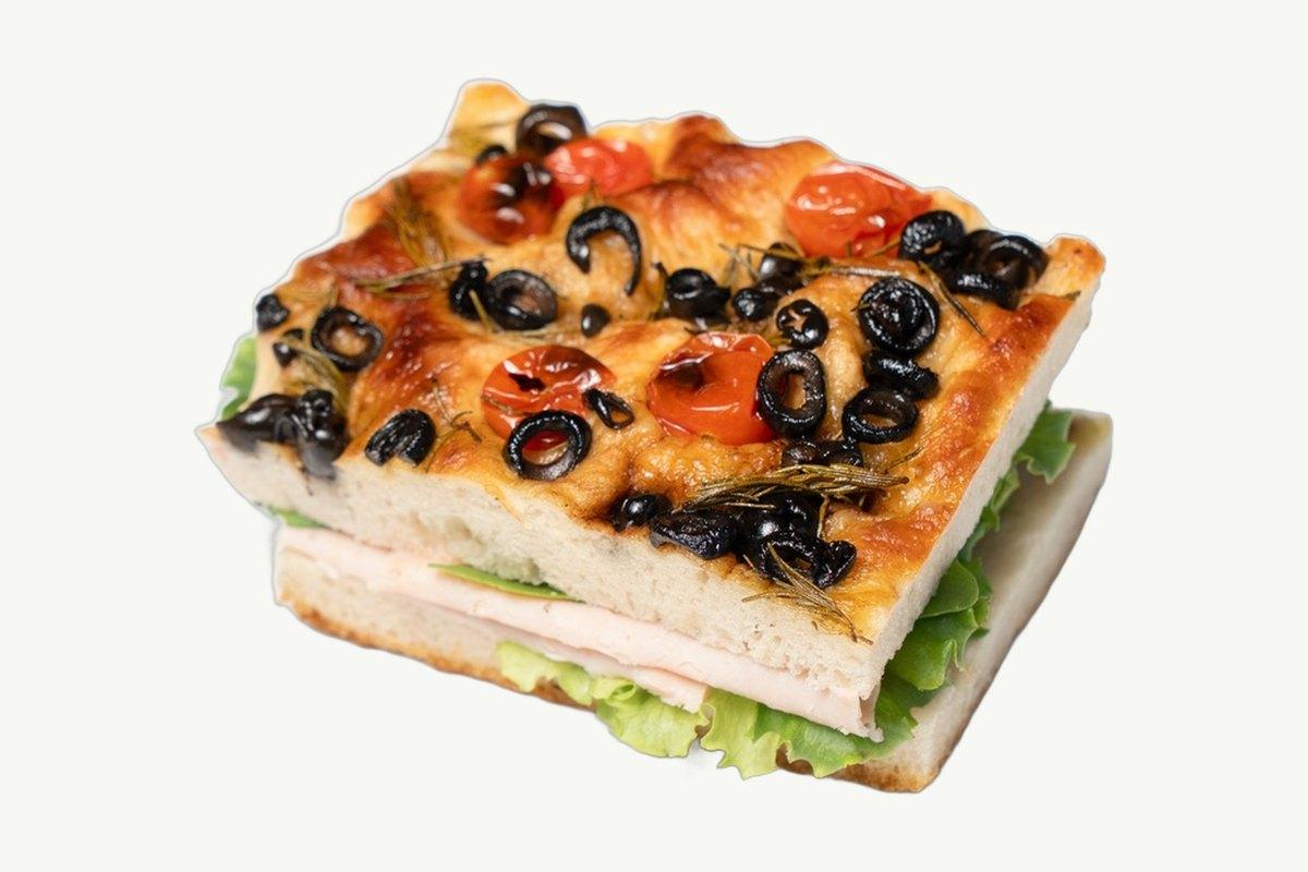 A square of focaccia topped with black olives and cherry tomatoes, filled with smoked turkey and lettuce, on a plain cream background