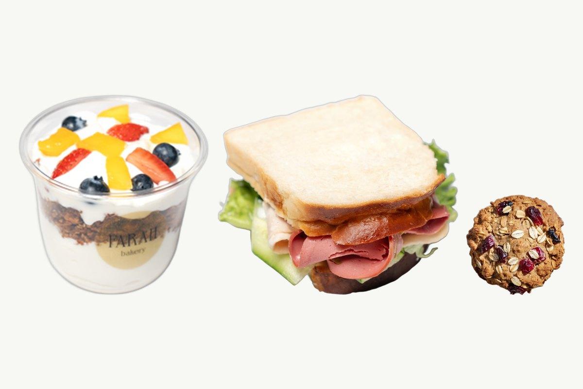 A granola parfait cup with fresh fruit, a white bread turkey sandwich and an oats and cranberry cookie on a plain cream background