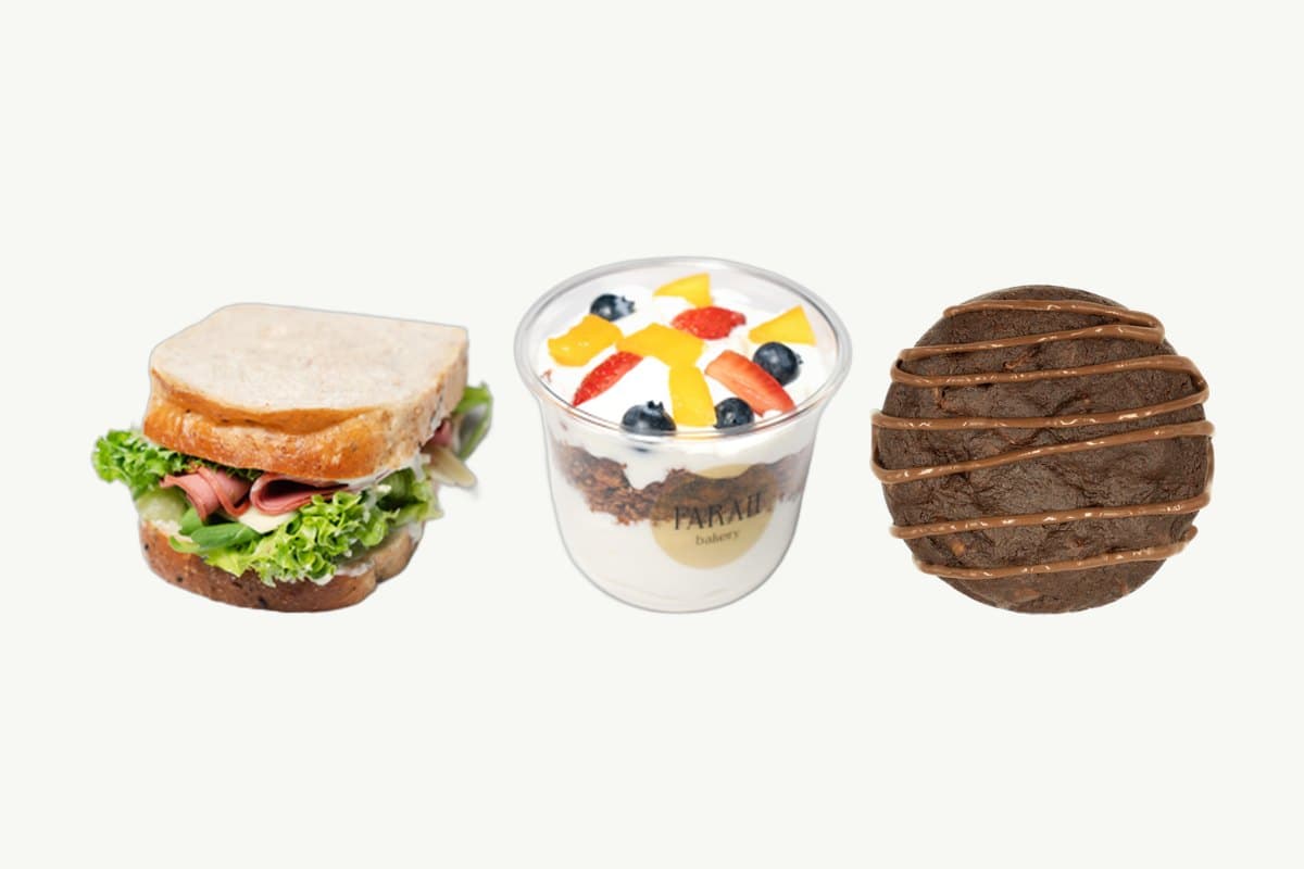 A multi-grain ham and cheese sandwich, a granola parfait with Greek yogurt and fresh fruit, and a double chocolate chip cookie in a row on a plain cream background