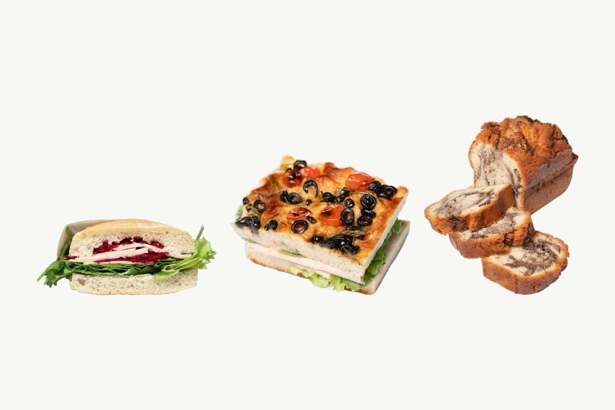 A ciabatta turkey and cheese sandwich, a focaccia smoked turkey sandwich topped with olives and cherry tomatoes, and a sliced marble cake loaf on a plain cream background