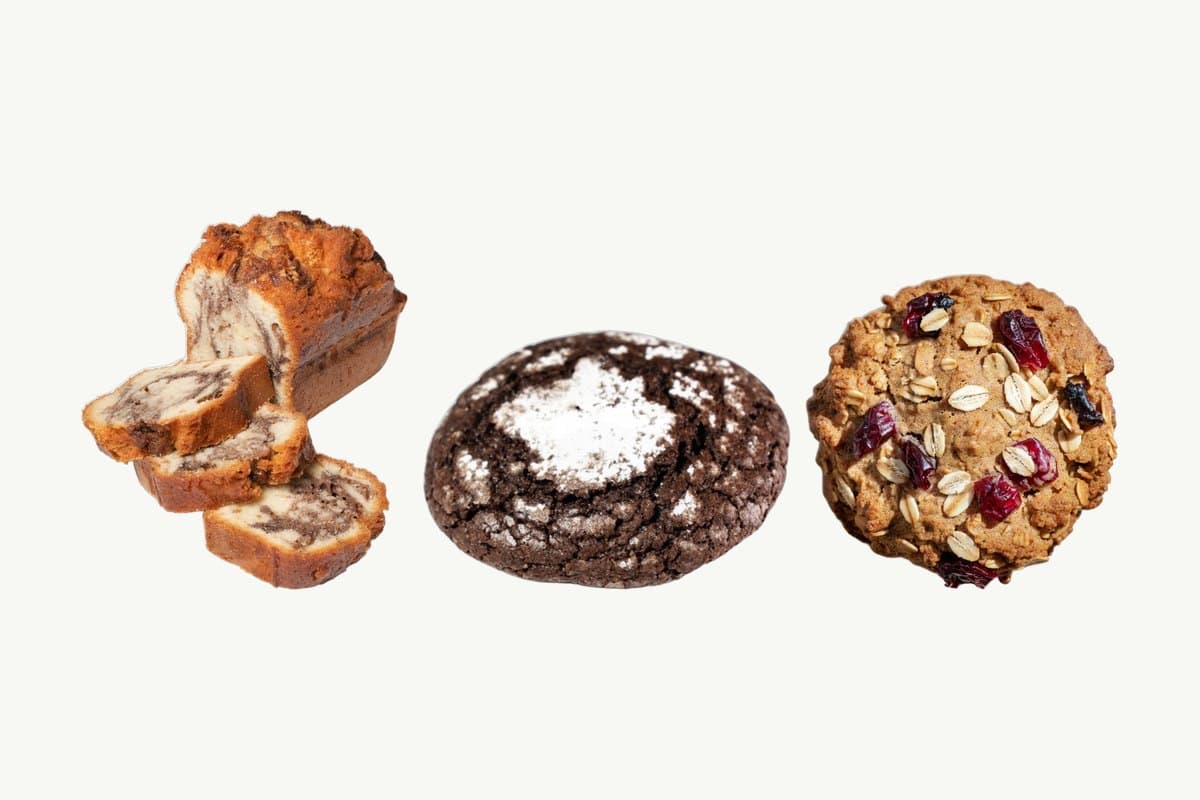 A sliced Marble Cake Loaf, a chocolate crinkle cookie and an oats and cranberry cookie in a row on a plain cream background