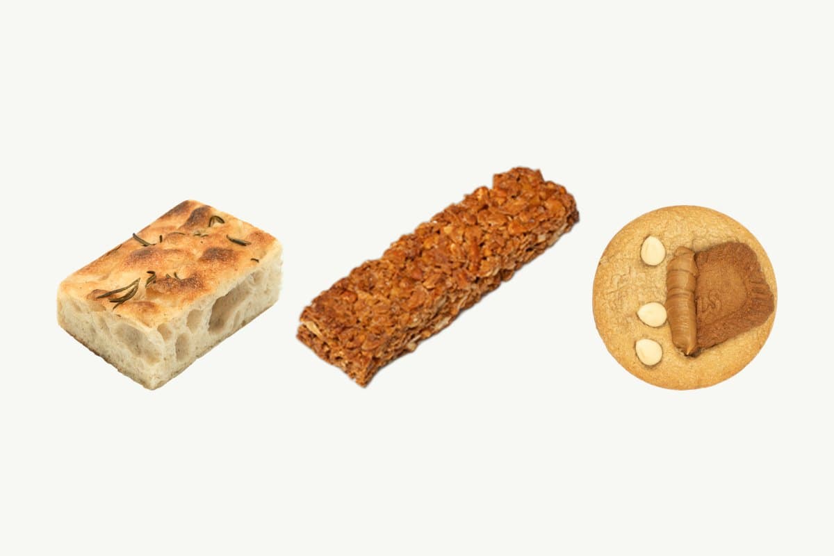 A rosemary and olive oil focaccia, a granola bar and a Lotus and white chocolate chip cookie in a row on a plain cream background