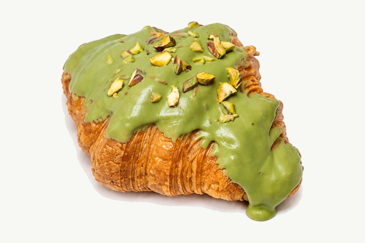 A croissant under a green glaze scattered with crushed pistachios, on a plain cream background