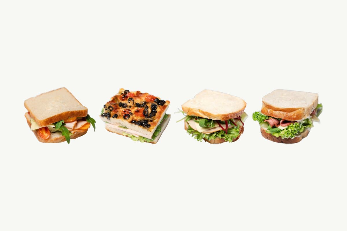 Four of our sandwiches, smoked chicken on white bread, smoked turkey on focaccia, smoked chicken on brioche, and ham and cheese, photographed on a plain cream background