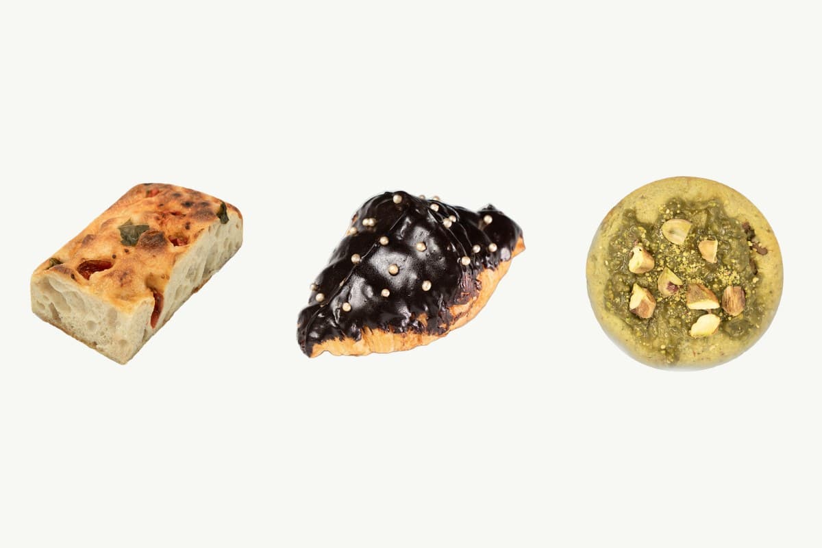 A cherry tomato focaccia slab, a chocolate croissant and a pistachio chocolate chip cookie on a plain cream background