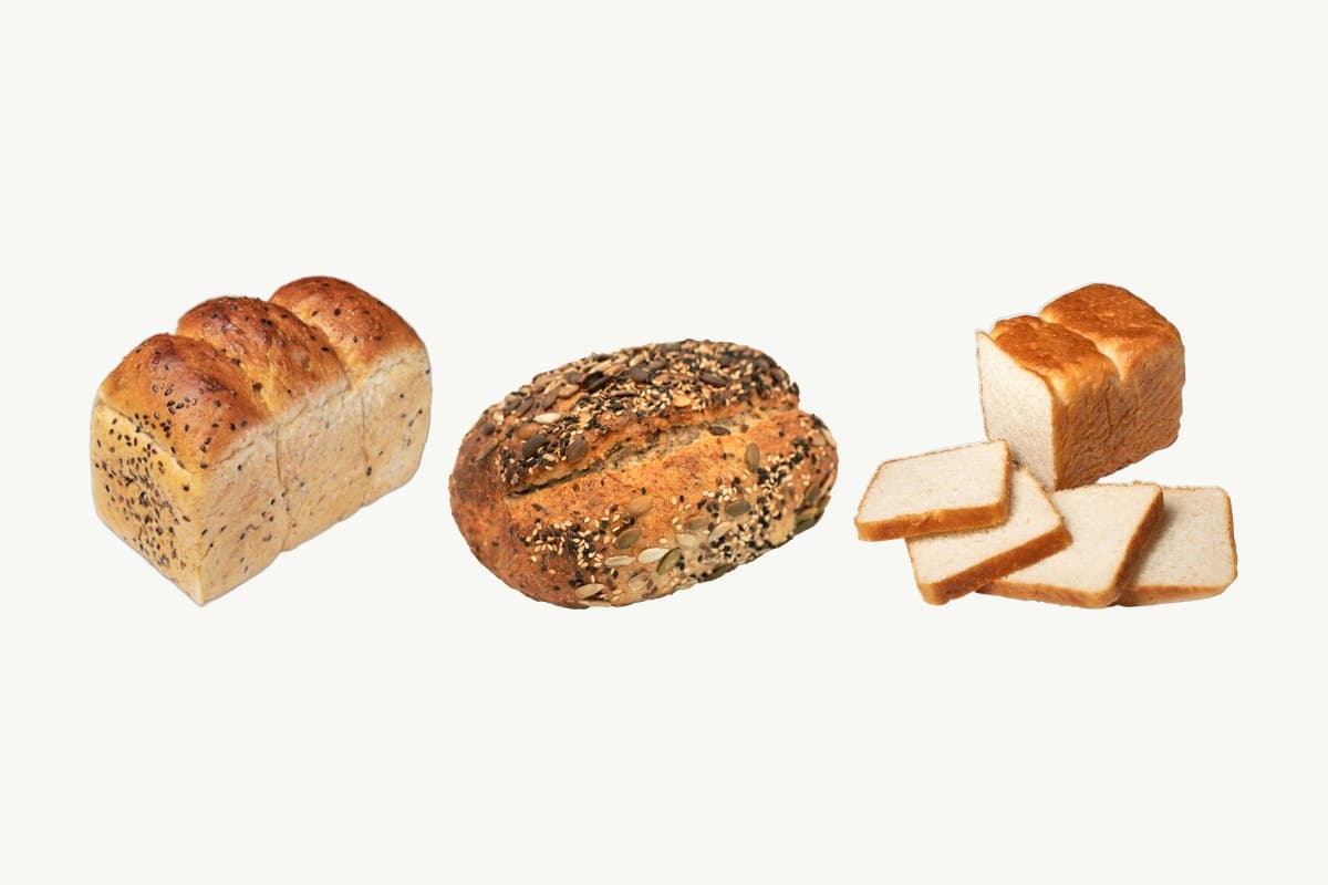 From left, a multi-seed whole wheat toast loaf, a multi-seed sourdough loaf and a sliced white toast loaf side by side on a plain cream background