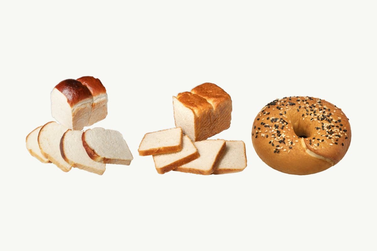 A brioche toast loaf, a sliced white toast loaf and a classic sesame bagel in a row on a plain cream background