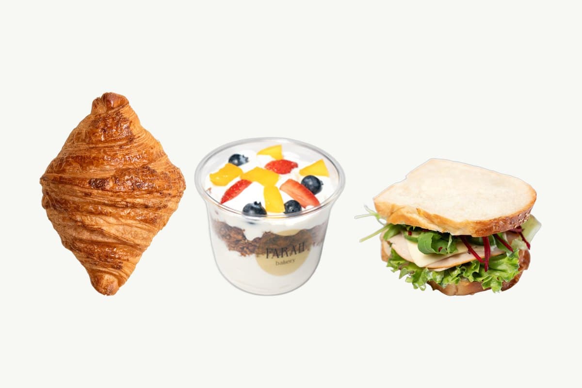A butter croissant, a Granola Parfait with Greek Yogurt in its cup and a brioche smoked chicken sandwich in a row on a plain cream background
