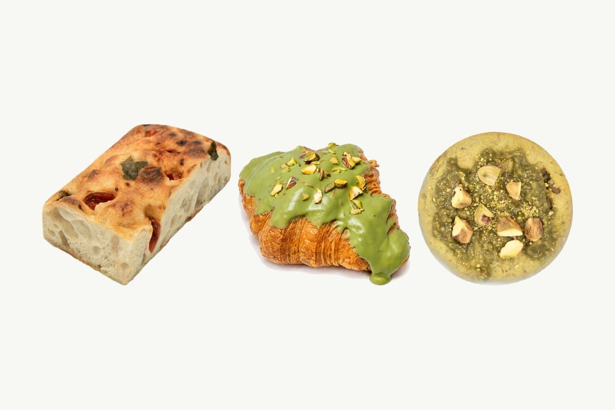 A piece of cherry tomato focaccia, a pistachio croissant and a pistachio chocolate chip cookie in a row on a plain cream background