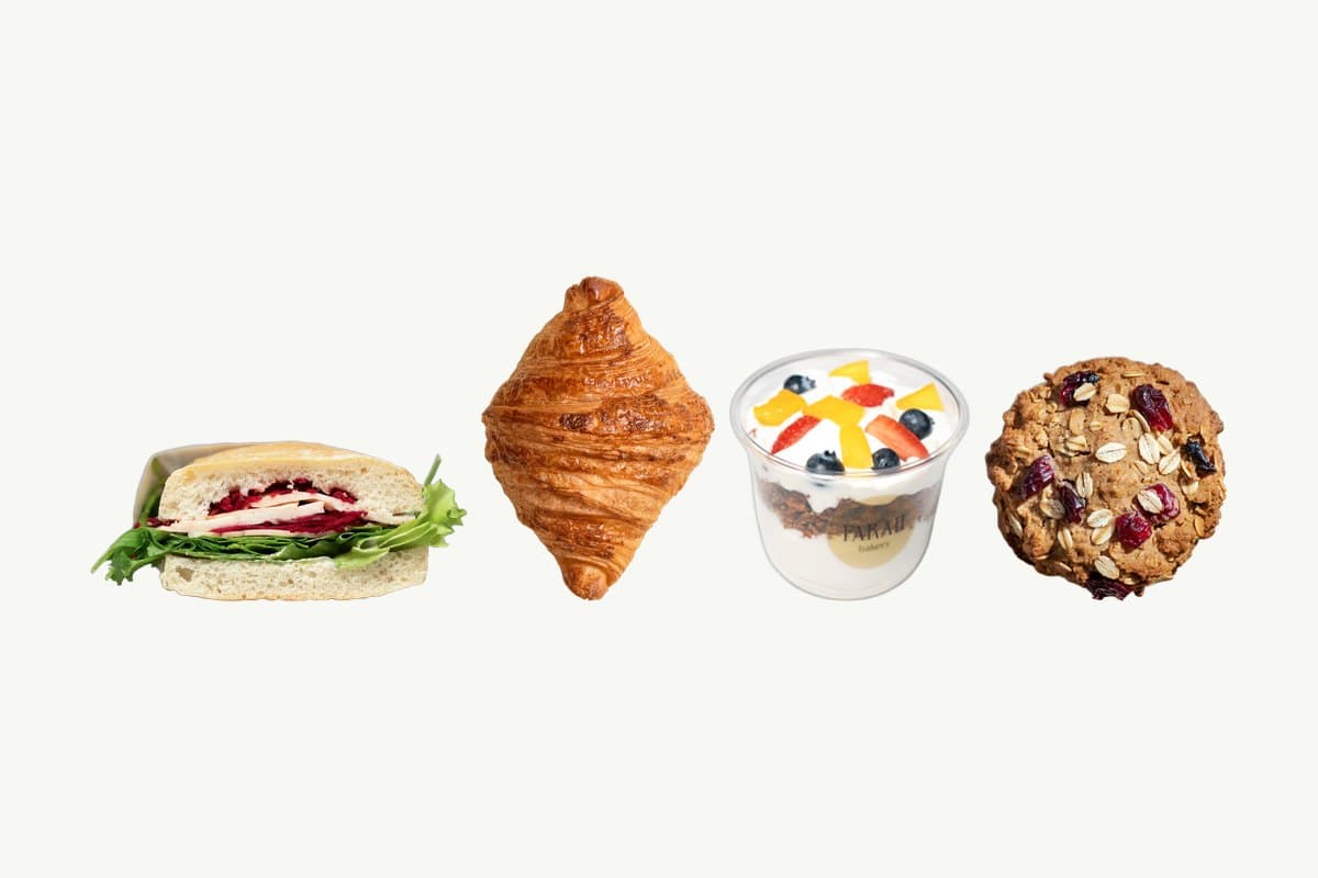 A ciabatta turkey and cheese sandwich, a plain croissant, a granola parfait with fresh fruit and an oats and cranberry cookie in a row on a plain cream background