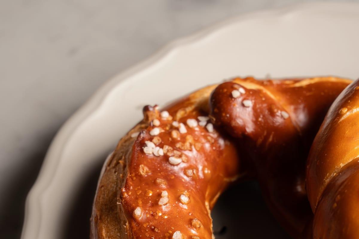 Glossy dark-browned salt-flecked pretzel resting on a cream scalloped plate against a moody background