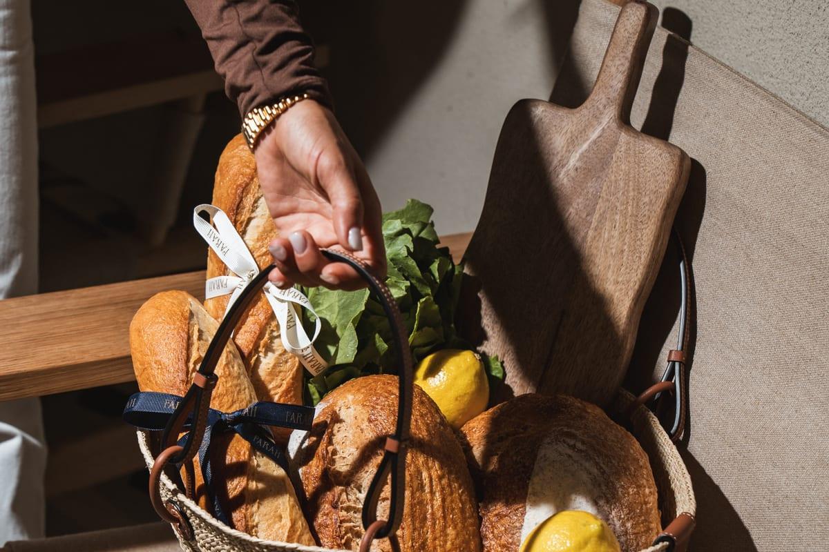 A hand lifting a woven basket of sourdough loaves, baguettes and lemons in warm sunlight