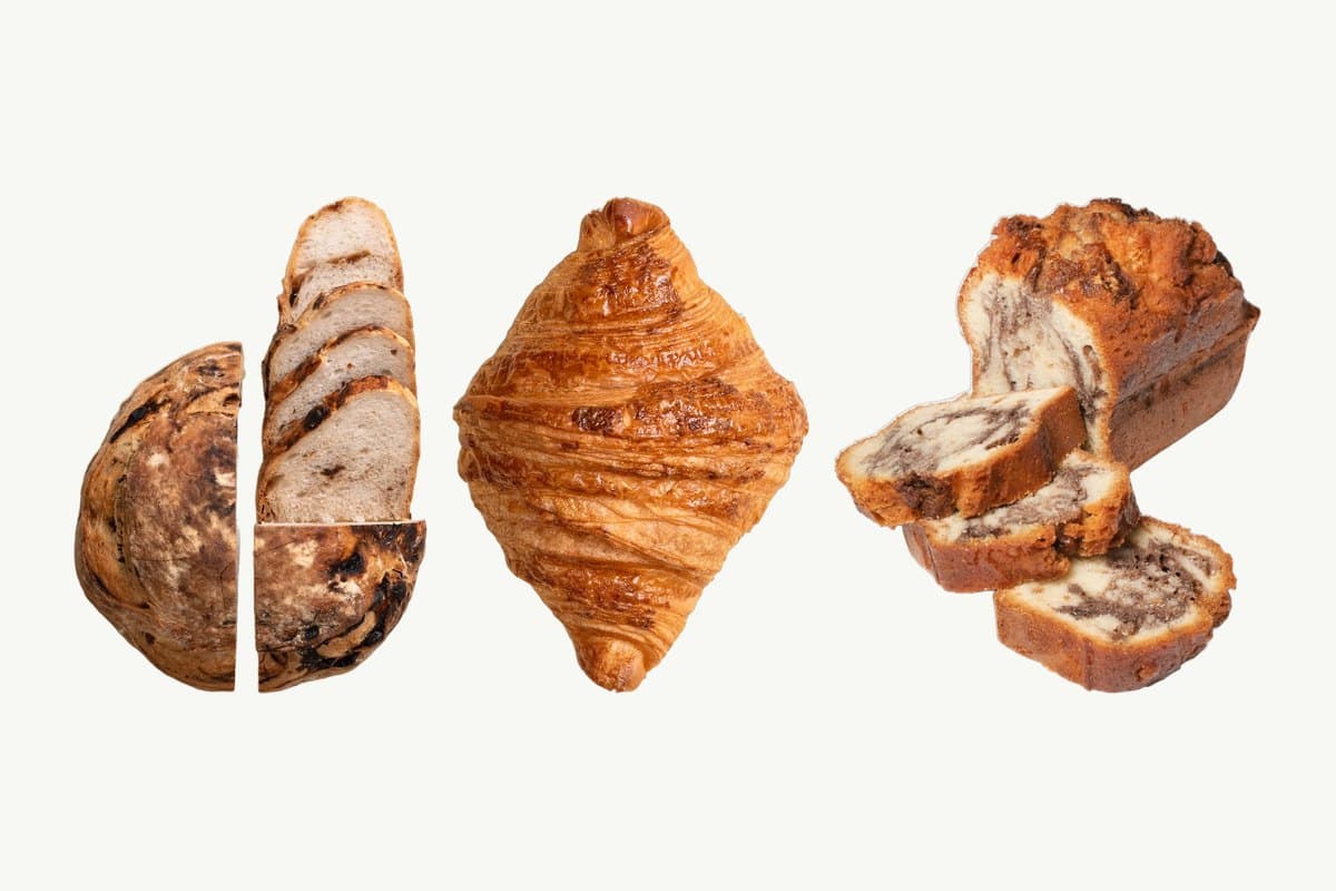 A Date, Walnut & Honey Sourdough loaf cut in half and sliced, a croissant, and a Marble Cake Loaf with two slices cut, on a plain cream background