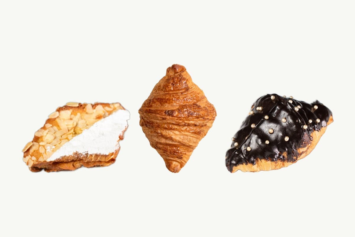 An almond cream croissant with toasted almonds and icing sugar, a plain butter croissant and a chocolate-coated croissant side by side on a plain cream background